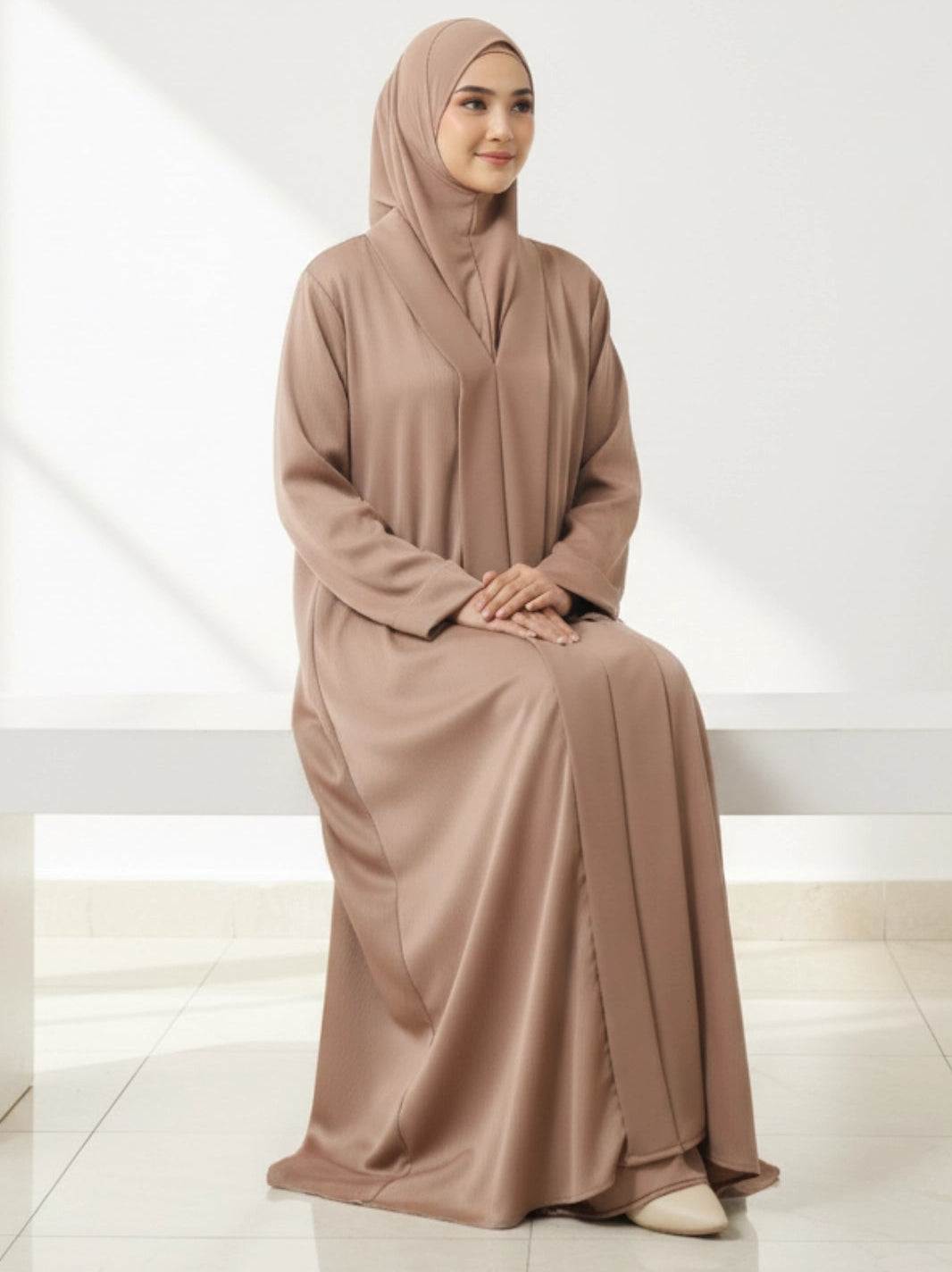 Woman wearing a brown abaya sitting on a white bench in a minimalistic setting
